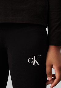 Svarta bomullsleggings har en vit stiliserad 'cK'-logotyp på vänster ben. Tyget är mjukt och elastiskt, lämpligt för vardagsbruk.
