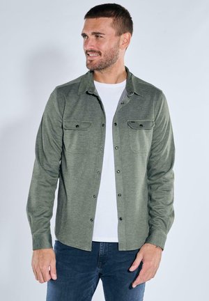 Grünes Langarmhemd mit Button-Down-Kragen, zwei Brusttaschen und subtiler Textur. Getragen über einem weißen T-Shirt. Normale Passform, lässiger Stil.