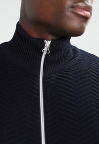 Strikket svart zip-up genser med høy krage, med ribbestruktur og chevronmønster, akkompagnert av en sølvglidert.