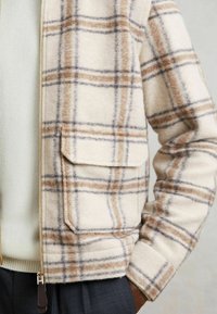 Beige ternet zip-jakke med stort klaplomme, båret over en creme sweater og parret med mørke bukser, med hånden delvist i lommen.