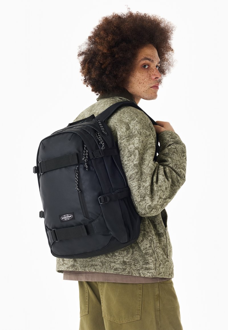 Jeune personne aux cheveux bouclés porte une veste verte à motifs et un grand sac à dos noir sur une épaule, regardant sur le côté.