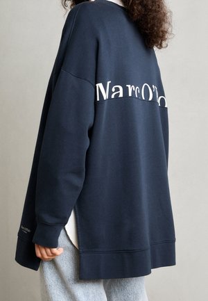 Person wearing oversized navy sweatshirt with white "Marc O'Polo" lettering across the upper back, paired with light gray pants.