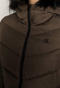 Brun pufferjacka med dragkedja, som har huva och chevronmönster. Broderad logotyp i svart nära dragkedjan. Slät textur.