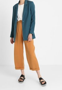 Blazer azul verdoso sobre camiseta blanca, combinado con pantalones de pierna ancha mostaza. Sandalias negras con suelas blancas. Texturas suaves y ajuste relajado.