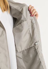 Vaaleanharmaa takki, jossa on sileä, kiiltävä kangas, sisätasku sekä neppari- ja nappikiinnitys kauluksessa ja hihoissa.