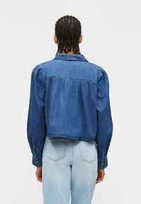 Άτομο με πλεγμένα μαλλιά φοράει μπλε τζιν μπουφάν μ ε φουσκωτά μακριά μανίκια και ανοιχτόχρωμο τζιν ψηλόμεσο, εμφανιζόμενο από πίσω.