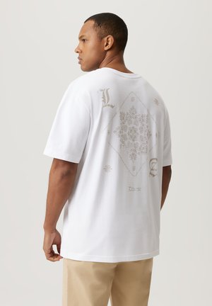 Biała koszulka z krótkim rękawem z dużym dekoracyjnym wzorem na plecach w jasnym beżu, złożonym z misternych wzorów i tekstu. Luźny krój.