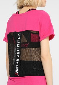 Růžový top s krátkým rukávem a černým síťovaným zadním dílem. Na boku je potisk "UNLIMITED BY HKMX" v bílé barvě, který ukazuje vrstvený design.