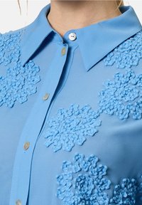 Chemise bleu clair à boutons avec col, ornée de broderies florales texturées dans un bleu plus foncé. Le tissu semble lisse et légèrement brillant.