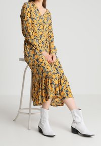 Gul blommönsterklänning med V-ringning och långa ärmar, med en volangslut. Matchad med vita cowboystövlar som når till ankeln.