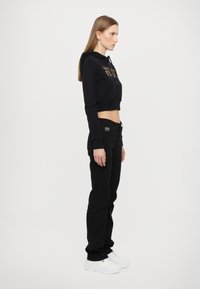 Zwarte cropped hoodie met gouden letters, gecombineerd met zwarte skinny jeans en witte sneakers, getoond vanuit een zijprofiel.