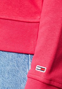 Primo piano di un polsino di felpa rossa con il piccolo logo Tommy Hilfiger sopra l'orlo a costine, abbinato a jeans di denim azzurri.