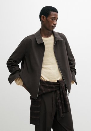 Mann trägt eine dunkelbraune Reißverschlussjacke über einem beigen Pullover mit einem karierten Hemd, das um die Taille gebunden ist, vor einem schlichten hellen Hintergrund.