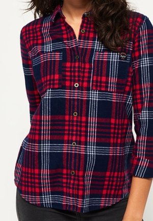 Vrouw draagt een geruit overhemd in rood, marineblauw en wit met twee borstzakken en opgerolde mouwen.