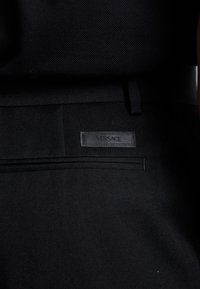 Mustat kangashousut, joissa on tekstuuri pinta, hienovarainen silmäys ja musta Versace-tunnus ommeltuna takataskuun.