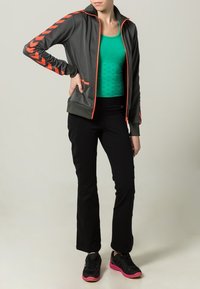 Grå zip-up jacka med orange detaljer och chevronmönster, lagd över en grön linne, matchad med svarta träningsbyxor och svarta sneakers.