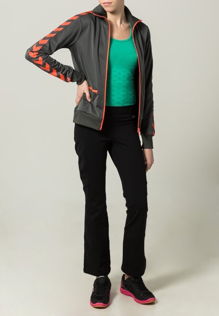 Grå zip-up jacka med orange detaljer och chevronmönster, lagd över en grön linne, matchad med svarta träningsbyxor och svarta sneakers.