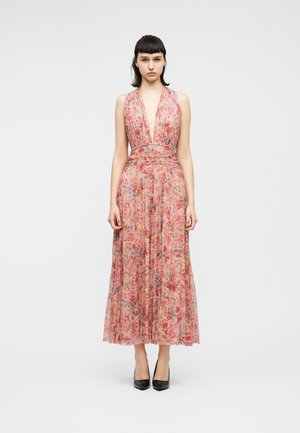 Žena v ružových tónoch, ktorá má na sebe bezrukávové kvetinové šaty s hlbokým výstrihom do V po členky, v kombinácii s čiernymi lodičkami na vysokom opätku, stojaca na neutrálom pozadí.