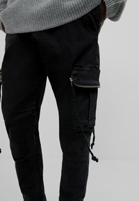 Svarta cargo-byxor med en matt textur, utrustade med en dragkedjeficka på låret, snörningsdetaljer och en slim fit-design.