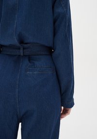 Džínový overal v tmavě modré barvě, se páskem v pase, dlouhými rukávy a zadní kapsou s elegantním prošíváním. Hladká, texturovaná tkanina.