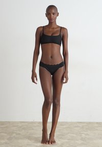 Čierny dvojdielny bikini plavky so zúženým topom s tenkými ramienkami a vlnitými okrajmi, doplnené o zodpovedajúce nízke nohavičky. Hladká textúra.