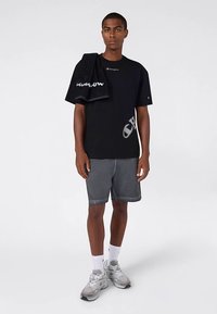 Zwart T-shirt met korte mouwen en logo, grijze versleten korte broek, witte sokken en grijze sneakers. Een zwarte handdoek over de schouder gedrapeerd.