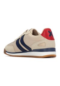 Zapatilla beis con rayas azul oscuro, suela de goma y acento rojo en el talón. Presenta una mezcla de materiales suaves y texturizados. Diseño de perfil bajo.