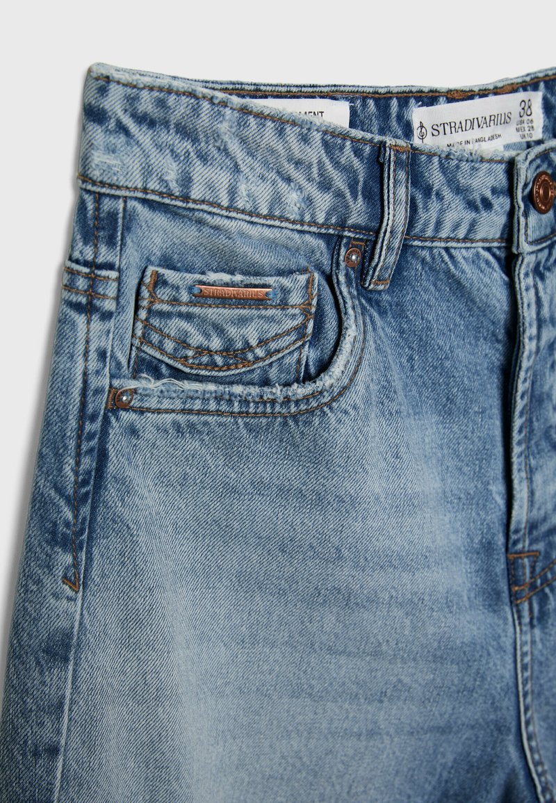 Jean en denim bleu clair avec une texture légèrement délavée, comportant des éléments en métal cuivré, une poche arrière et une étiquette à l'intérieur de la ceinture.