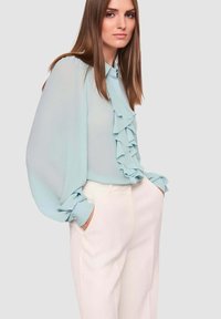 Blusa azzurro chiaro con davanti con volant, maniche lunghe a sbuffo e polsini con bottoni, abbinata a pantaloni bianchi sartoriali. Tessuto liscio e semitrasparente.