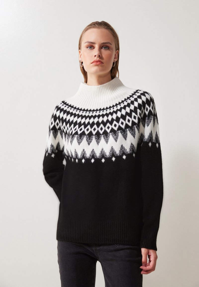 Svart og hvitt strikket genser med høy ribbestrikket krage og geometrisk fair isle-mønster over brystet, med en myk tekstur.