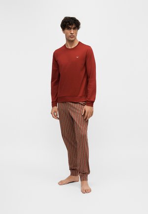 Homme portant une chemise rouge à manches longues et un pantalon rayé rouge, blanc et bleu, debout pieds nus sur un fond blanc uni.