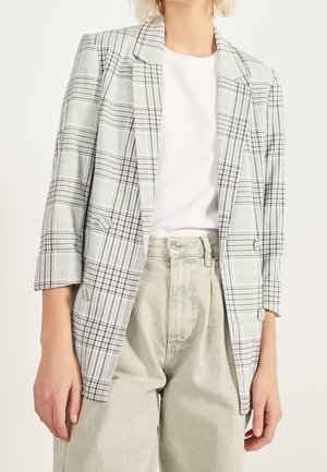 Personne portant un blazer à carreaux gris clair sur un t-shirt blanc et un pantalon beige taille haute, debout devant un fond uni.