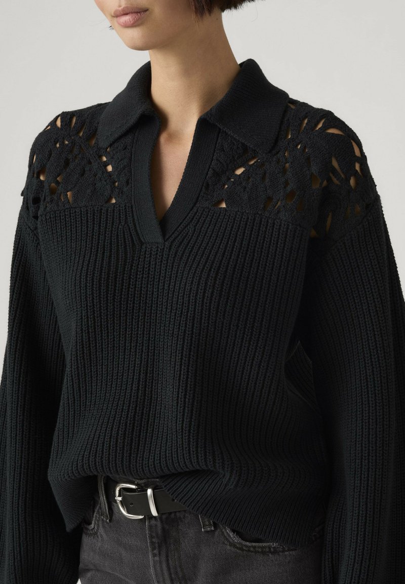 Vrouw draagt een zwarte gebreide trui met kantopeningen op de schouders en een kraag, gecombineerd met een zwarte riem en donkere spijkerbroek.