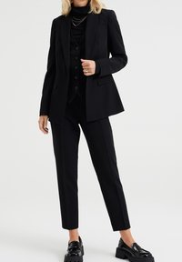 Svart skräddarsydd kostym med en kavaj med fickor och slag, parat med en figursydd väst och smala byxor. Glansiga svarta loafers.