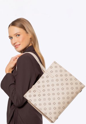 Žena s dlouhými blond vlasy, oblečená v tmavé bundě, nese na rameni béžovou tašku s ozdobnými vzory a mírně se usmívá.