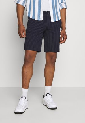 Bermuda a righe blu navy con vestibilità slim, dotate di chiusura a bottone e tasche, abbinate a sneakers bianche e una camicia a righe azzurra chiara.