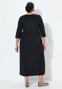 Musta, nilkkapituinen mekko, jossa on kolmesadelman hihat ja kerrottu vyötärö. Kangas on sileä. Yhdistetty teksturoituihin mustiin liukupolkimiin.