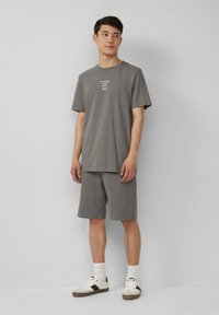 Graues Baumwoll-T-Shirt mit kurzen Ärmeln und Rundhalsausschnitt, kombiniert mit passenden grauen Shorts. Auffälliger Grafiktext auf der Vorderseite des Shirts. Weiße Sneakers.