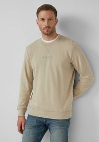 Béžová mikina s okrúhlym výstrihom vyrobená z textúrovanej látky, s pružnými manžetami a spodným lemom. Obsahuje decentné vyšívané logo na hrudi. Nosená s modrými džínsami.