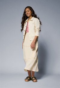 Beige denim jacket, light pink top, and cream skirt with texture; olive green sandals with bows; model has long wavy hair.