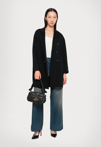 Schwarzer doppelt dekorierter Mantel mit großen Knöpfen, weißes Oberteil, hellblaue weite Jeans und eine gesteppte schwarze Handtasche mit goldenen Akzenten.