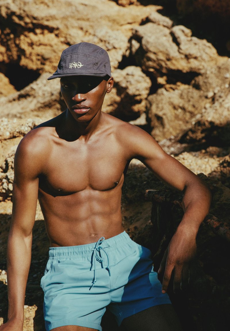 Homme torse nu portant une casquette bleu marine et un short de bain bleu clair, assis sur un terrain rocheux sous le soleil avec des ombres sur son visage et son corps.