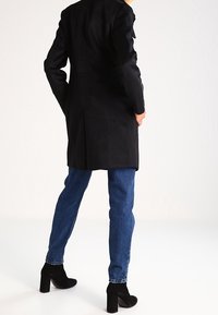 Cappotto lungo nero con design dritto, texture liscia, cuciture dettagliate, abbinato a jeans in denim blu e stivali neri alla caviglia.