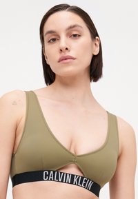 Olijfgroene bralette met een driehoekig ontwerp, met een uitsnijding en een zwarte elastische band met het "CALVIN KLEIN" logo. Gladde textuur.