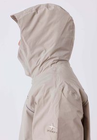 Chaqueta con capucha de color beige claro con una textura suave, que cuenta con un logo en el hombro y costuras de acento sutiles a lo largo de las costuras.