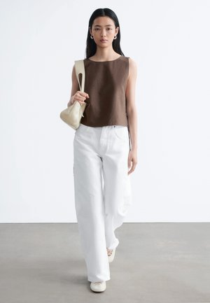 Femme marchant vers l'avant portant un haut marron sans manches, un pantalon blanc large, des chaussures crème et tenant un petit sac à bandoulière beige.