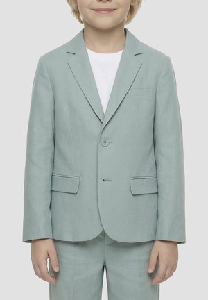 Kind trägt eine hellgrüne Sakko und passende Hose über einem weißen Hemd, steht vor einem einfarbigen Hintergrund.