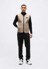 Νέος άνδρας στέκεται μπροστά φοράει ένα μπεζ καπιτονέ γιλέκο πάνω από μαύρο πουκάμισο με μακριά μανίκια, μαύρο παντελόνι και λευκά αθλητικά παπούτσια με μαύρες λεπτομέρειες.