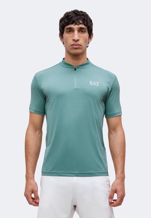Man draagt een teal kortemouwen shirt met rits en EA7-logo en witte short, kijkt recht vooruit tegen een effen lichte achtergrond.