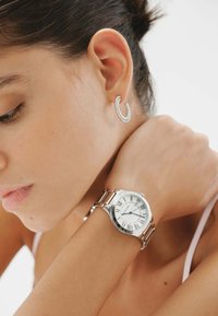 Reloj plateado con números romanos y manecillas azules en la muñeca, combinado con un pendiente de aro plateado texturizado. Piel suave visible en primer plano.